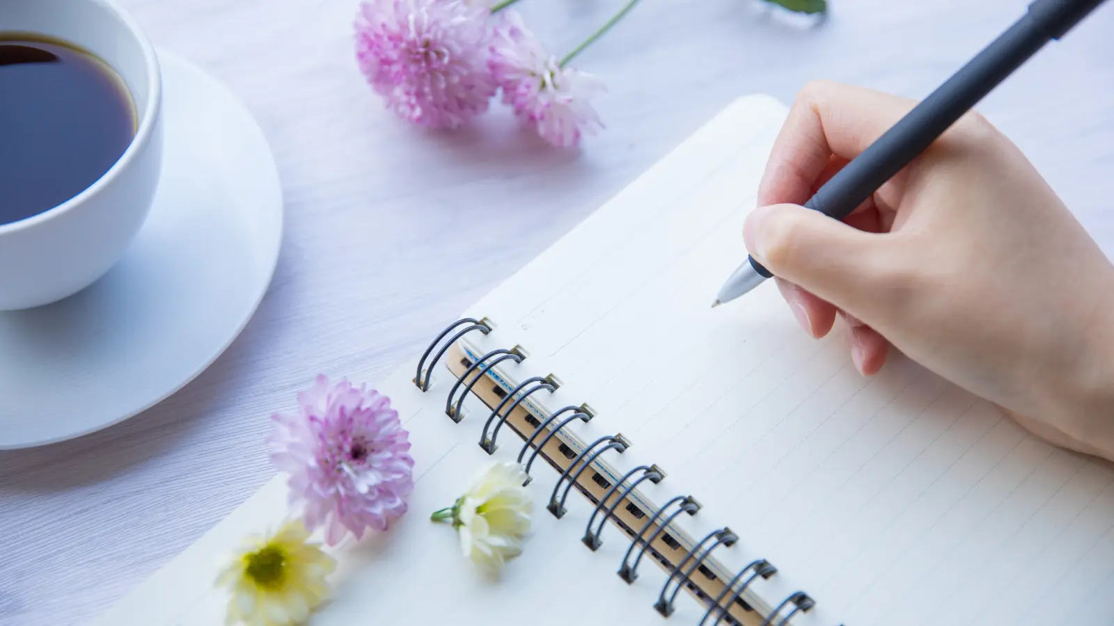 Una mano escribiendo sobre una libreta. En la mesa hay una taza y flores