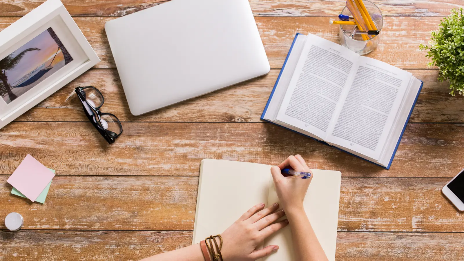 Una manos escribiendo en una libreta sobre una mesa de madera, en la que también hay dos libros abiertos